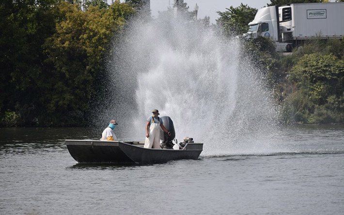 Two people wearing protective cloth face masks on a boat with a large outboard boat motor. One sits at the front of the boat while the other at the back of the boat steers, a large spary of water jets up into the sky as the motor is turned.